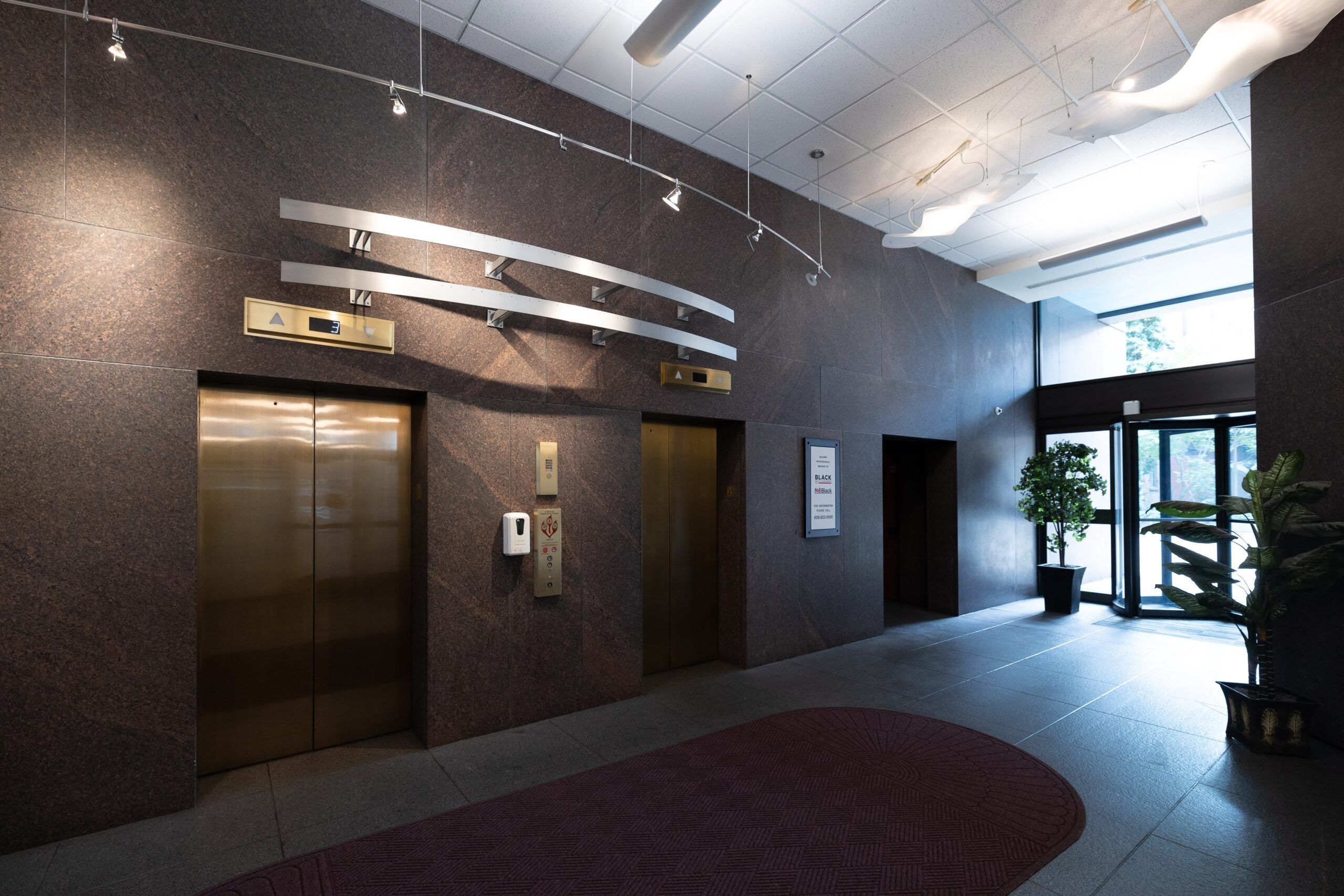 A lobby area with two closed elevator doors on the left, featuring metallic panels, highlights the quality of Commercial Elevator Services. The walls are clad in dark stone tiles. There's a red mat on the floor and a door to the outside at the back right, flanked by potted plants. Ceiling lights illuminate the area.