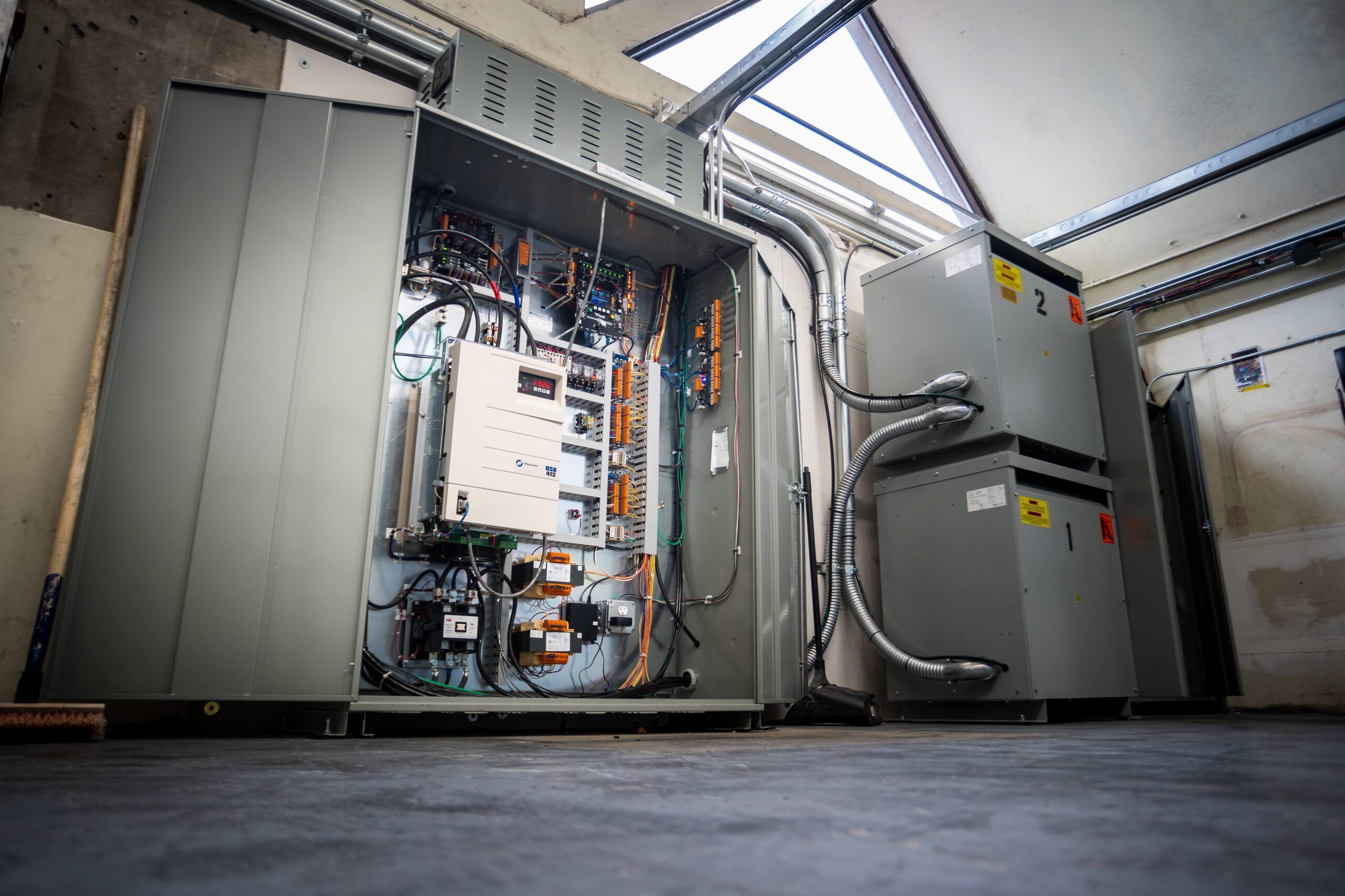 A room with electrical equipment. An open electrical cabinet on the left exposes wiring, circuit boards, and components. Adjacent to it are two large, gray electrical units with cables extending from them. The room has a concrete floor and partially sloped ceiling, ideal for an elevator apprenticeship program.