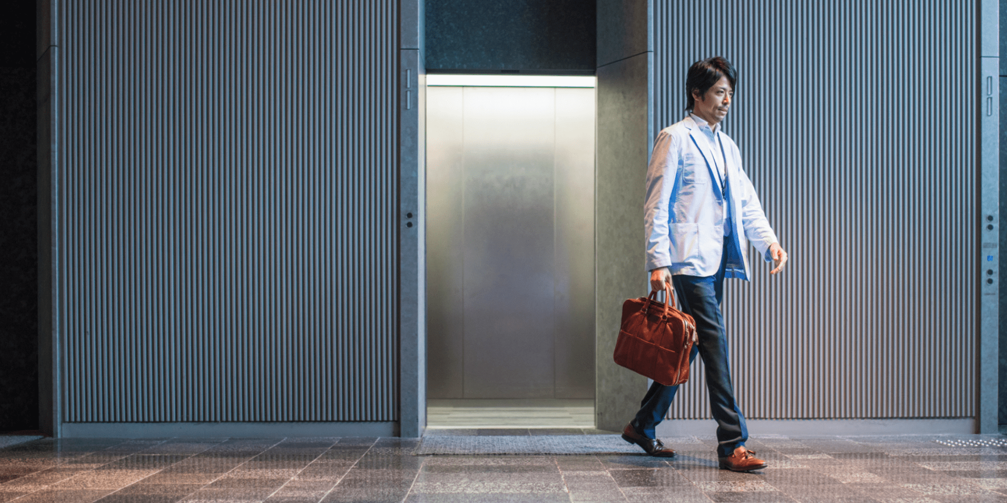 A person in business attire carrying a brown leather bag is walking away from an open home elevator. The individual is dressed in a light blue blazer, dark pants, and brown shoes. The surroundings include gray walls and a tiled floor.