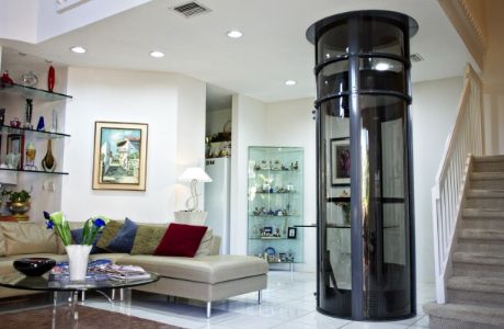 A modern living room featuring a transparent, home elevator in a cylindrical design. The room has a sectional beige sofa with various colored pillows, a glass coffee table, and shelves displaying decorative items. A staircase is visible on the right-hand side.
