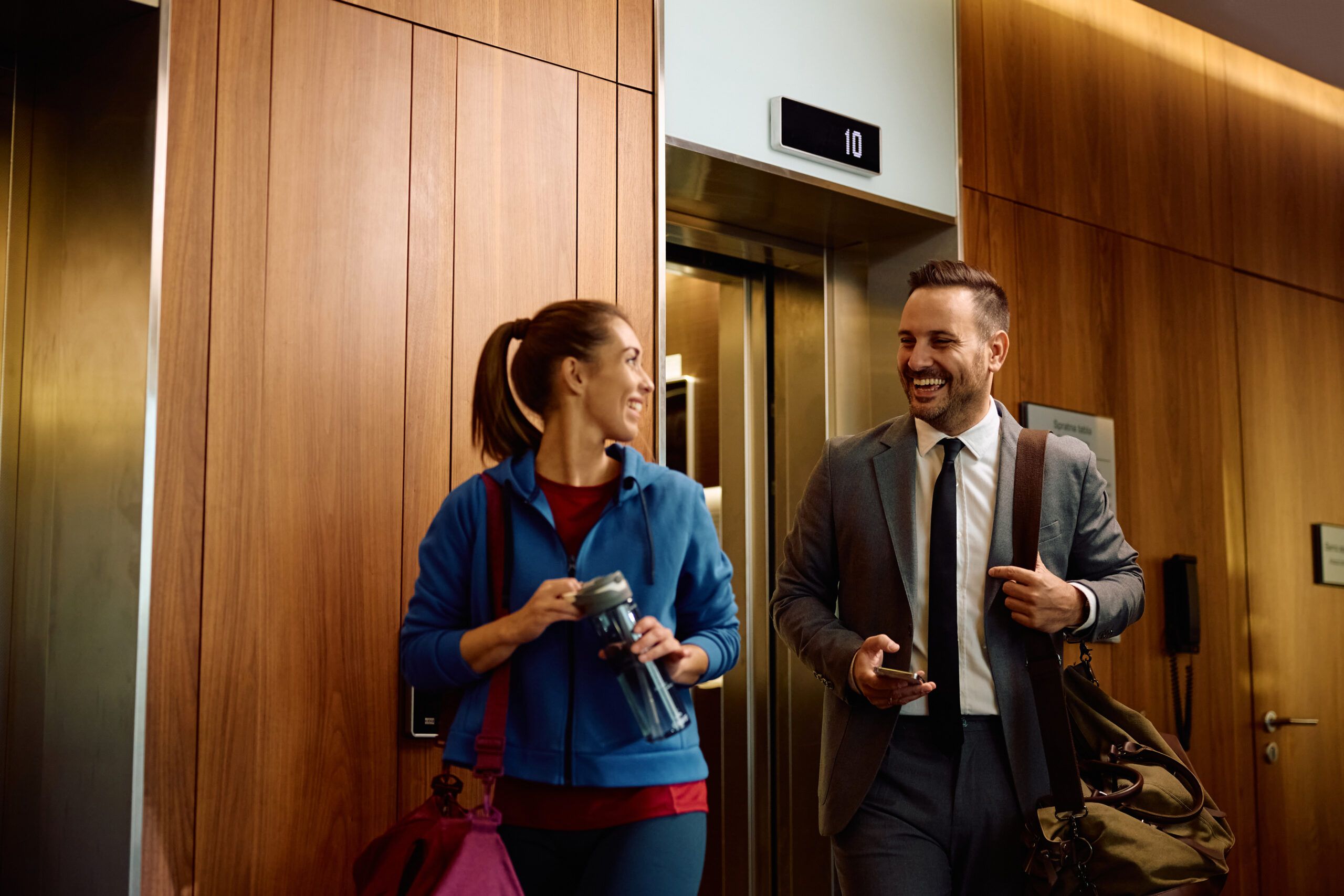 A woman in sportswear carrying a water bottle and gym bag walks beside a man in a suit holding a phone and briefcase. They pass by signs for elevator repair services in the hallway, illuminated by the lighting overhead against wooden panel walls.