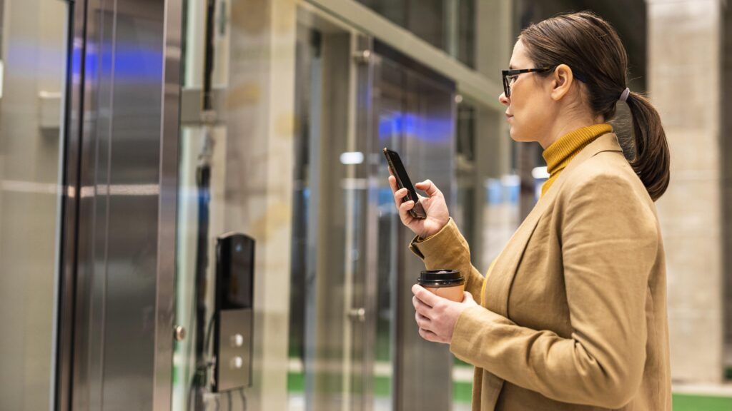 A woman wearing glasses and a tan blazer holds a coffee cup and looks at her smartphone while standing in front of glass doors in a modern building.